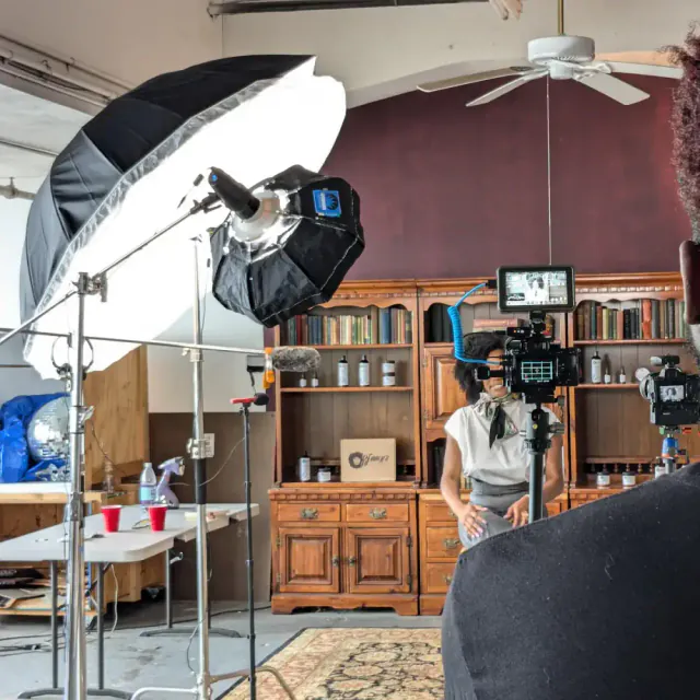 Camera, lighting and a monitor arranged to film a woman in front of product shelves.