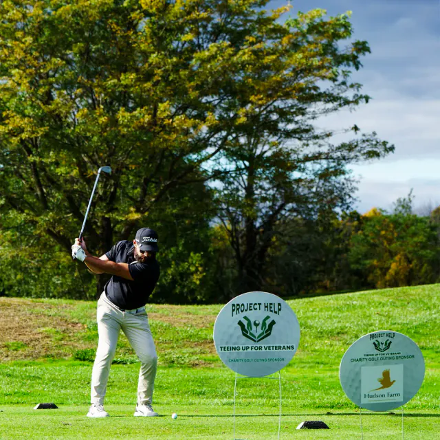 A golfer mid-swing beside Project Help “Teeing Up for Veterans” charity golf outing signs.